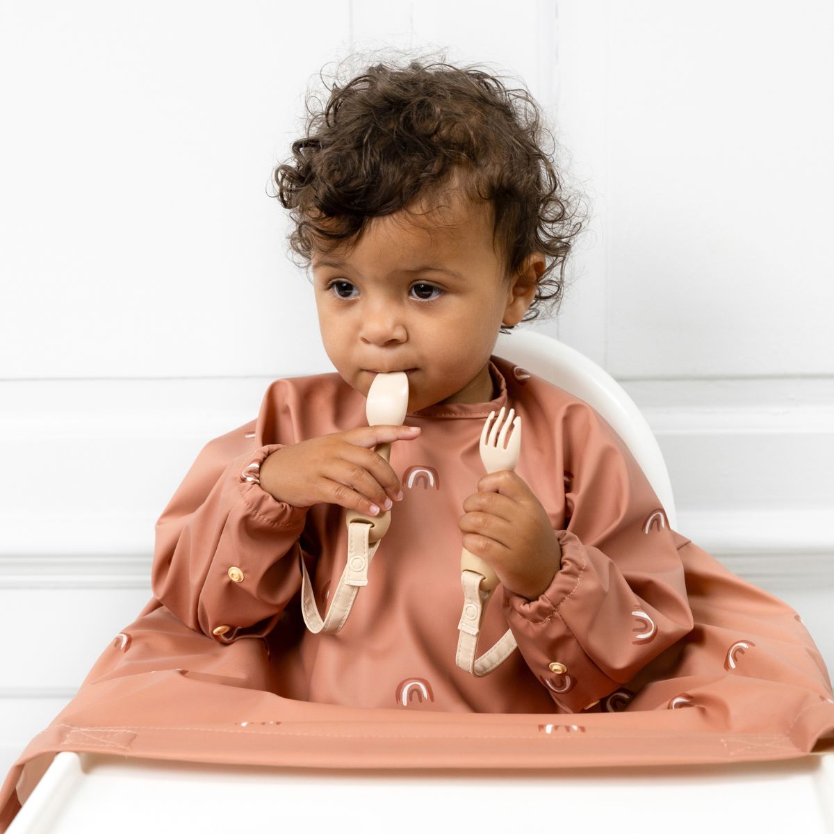 A baby feeding themselves with Bibado cutlery, wearing a Coverall attached to the highchair tray